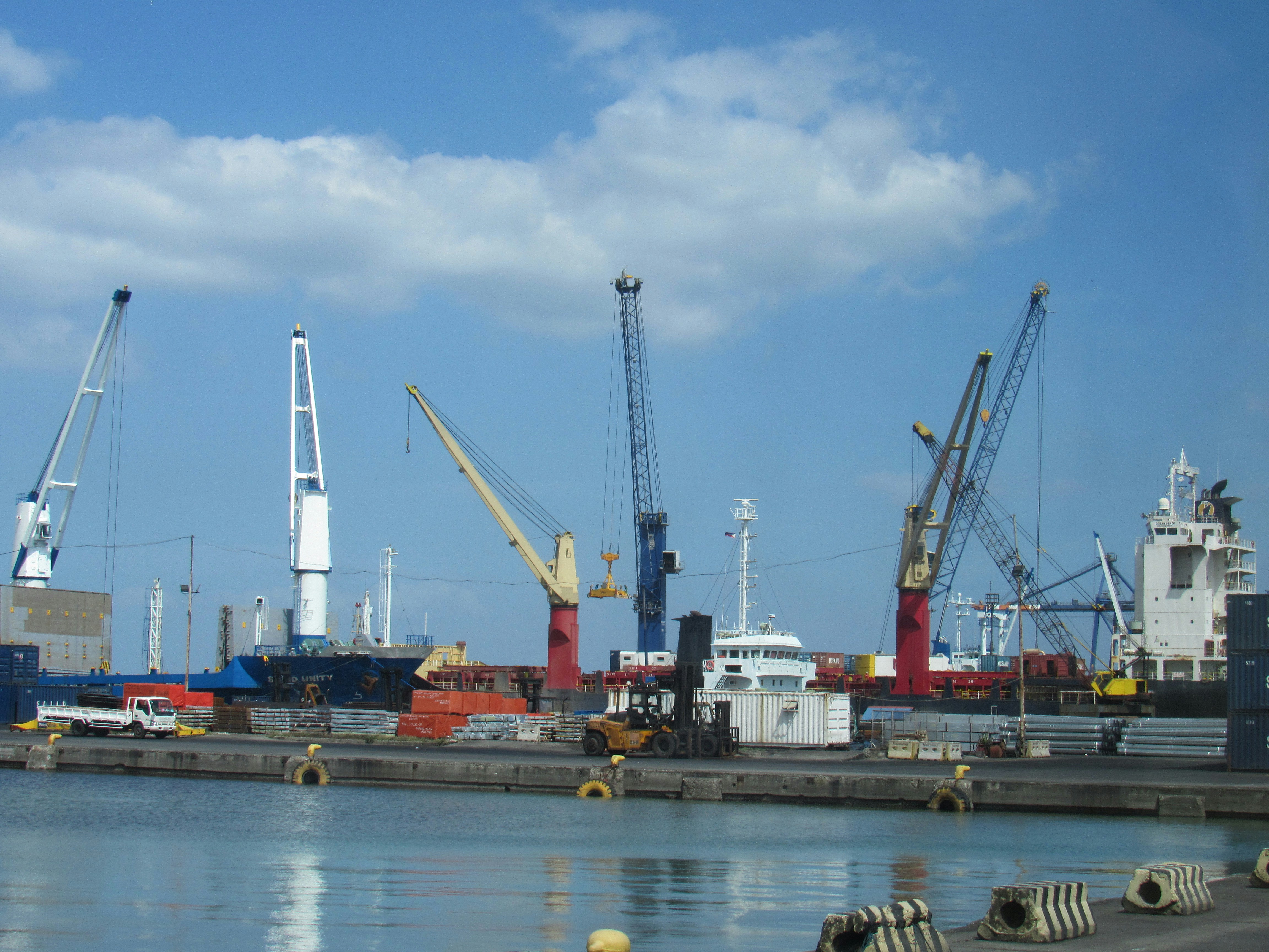 A busy harbour with several large ship cranes, cargo vessels, stacked containers and a forklift along the quayside under a blue sky.