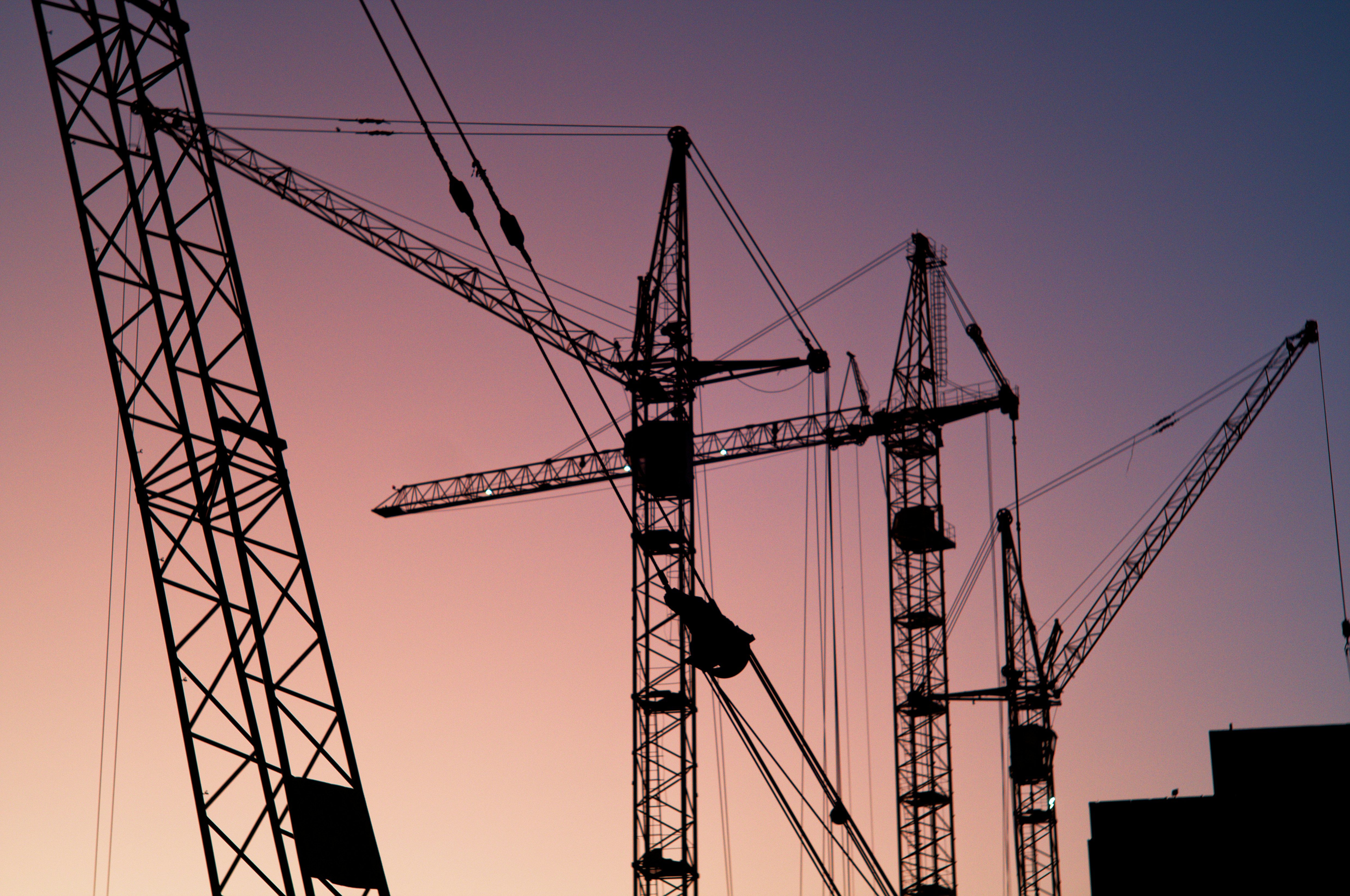 Several tower cranes are silhouetted against a pink and purple sunset sky above a construction site.