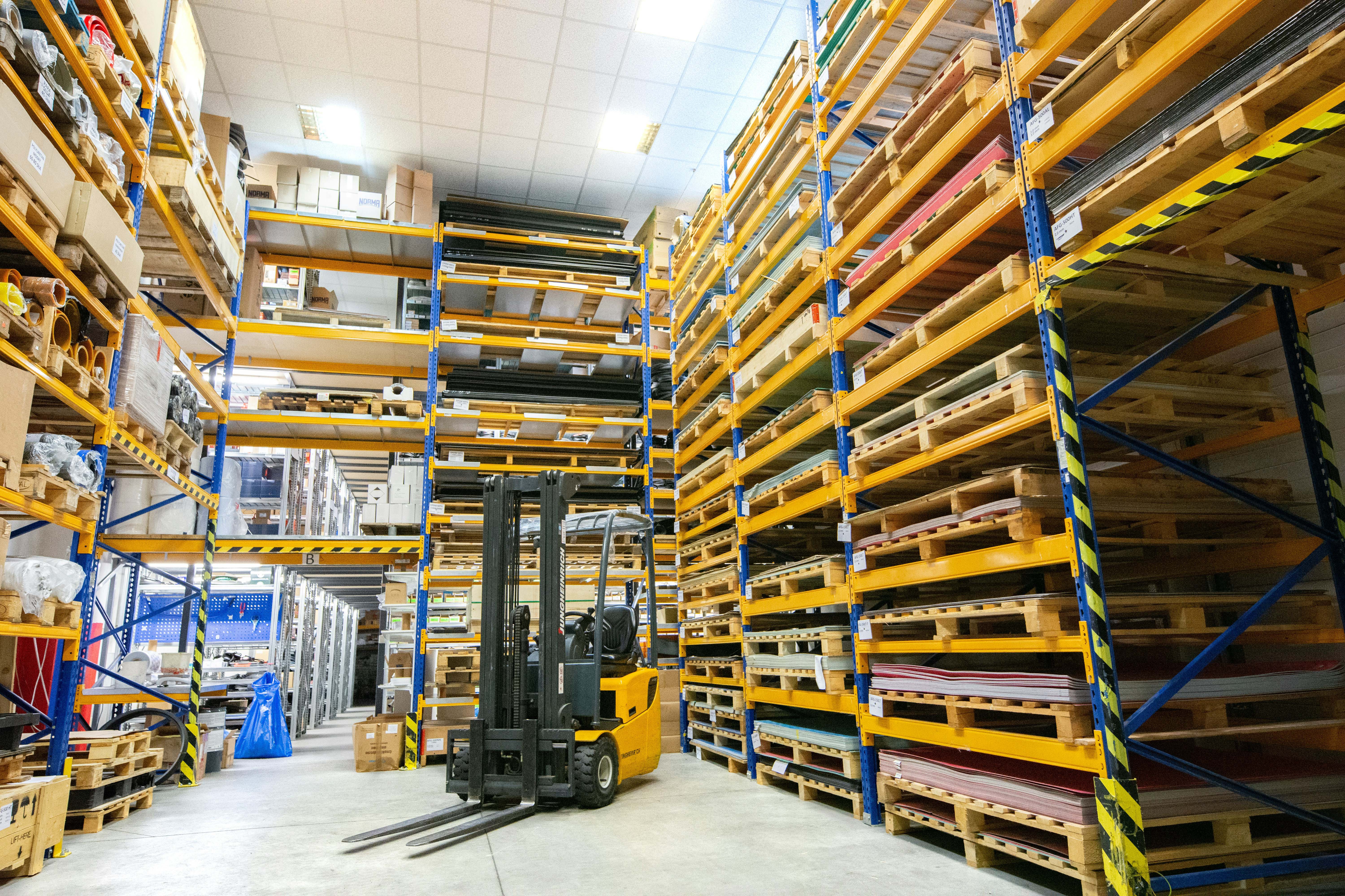A small unmanned forklift truck stands parked in a warehouse aisle lined with tall yellow and blue pallet racking stacked with boxes and sheet materials.
